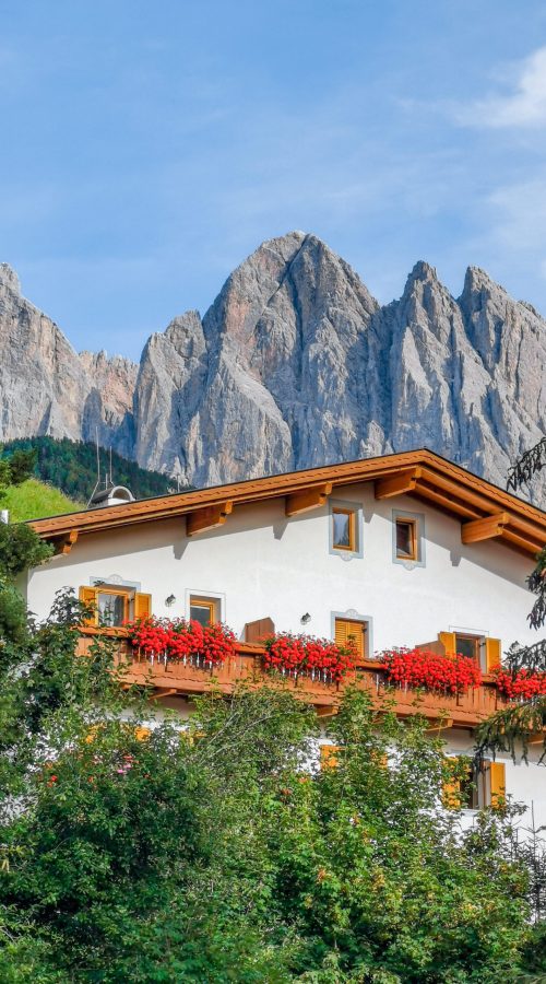 Gruppo delle Odle: cielo azzurro, casa di legno, fiori rossi nel davanzale, montagne sullo sfondo