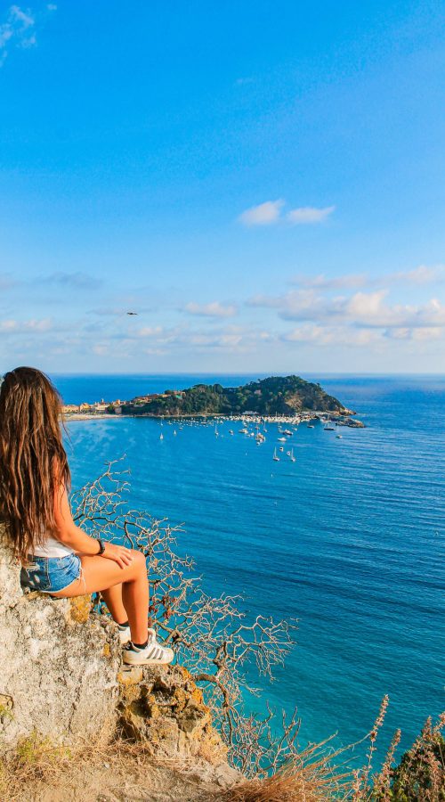 ragazza seduta su delle rocce con affaccio sul mare, cielo azzurro